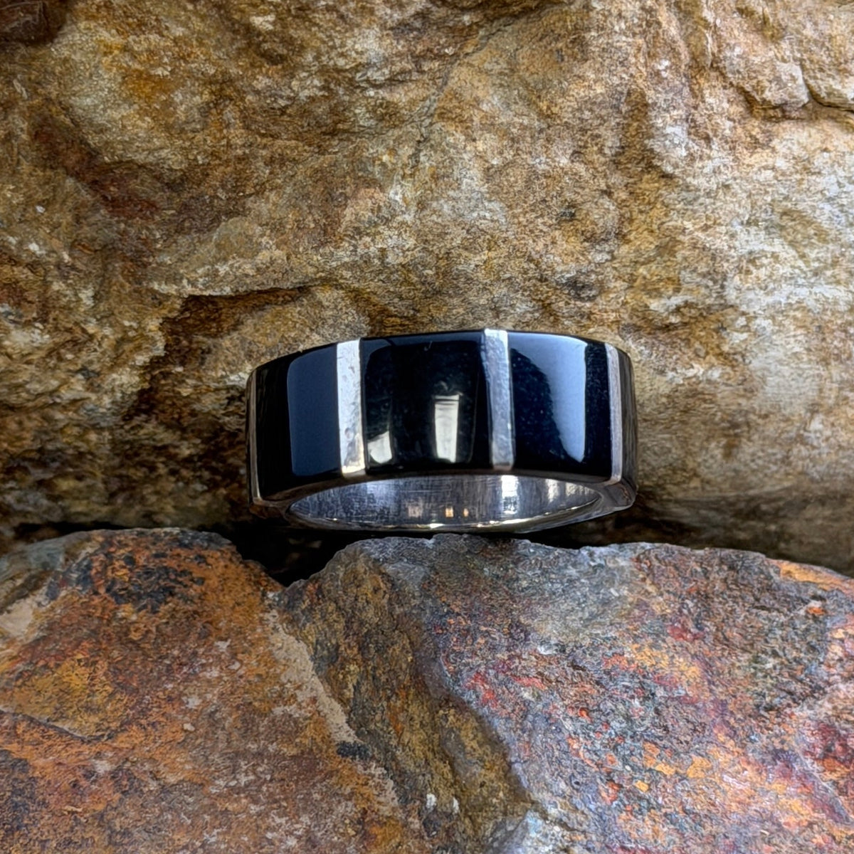 A polished black and silver banded ring resting on a textured orange-brown rock with a natural stone wall in the background.