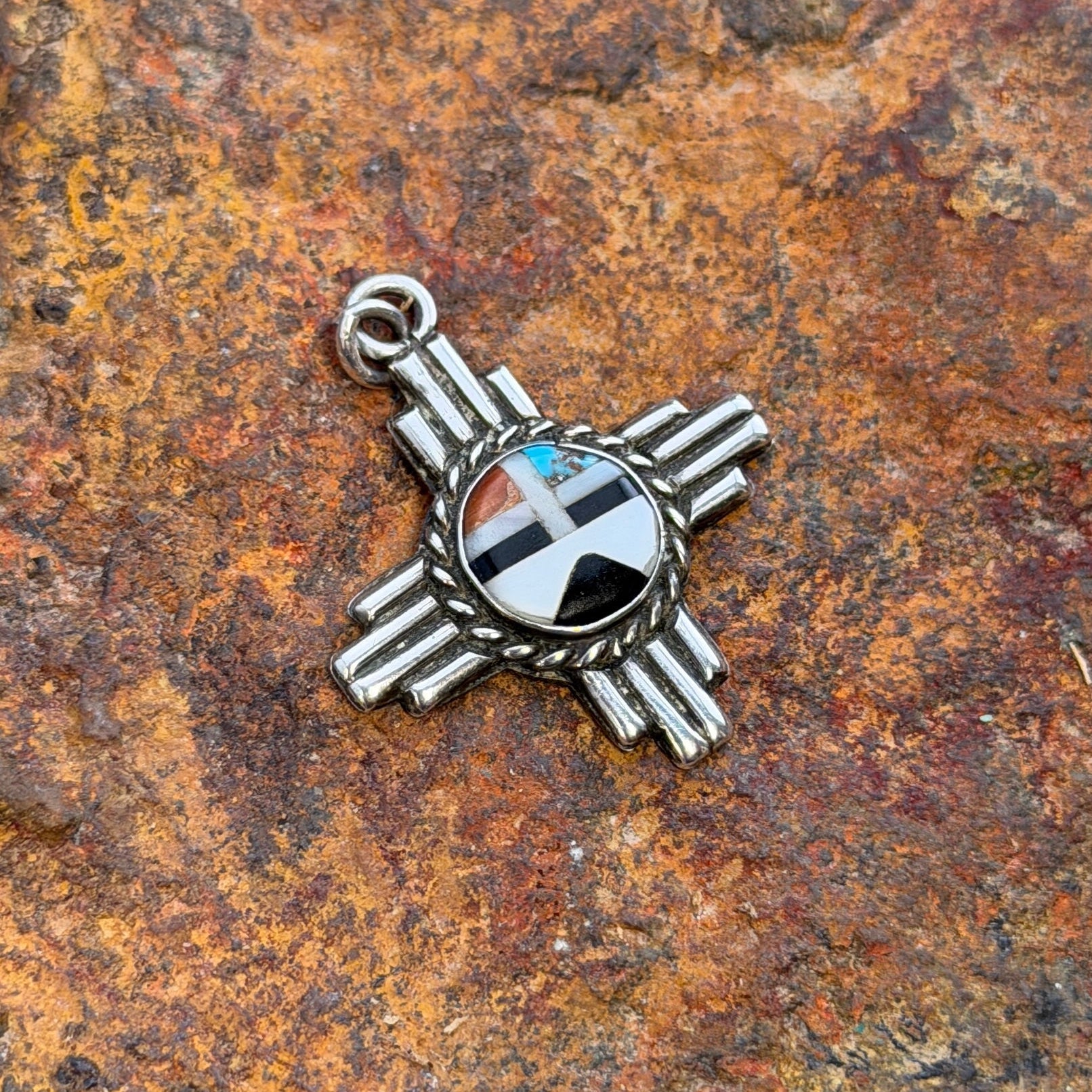 A silver-toned Zuni cross pendant with a circular inlay of black, white, red, and turquoise, displayed on a textured orange-brown rock.