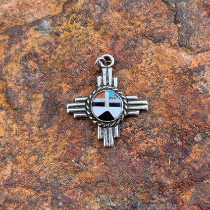 A silver-toned Zuni-style cross pendant with a black, white, and blue inlay design, displayed on a textured brown and orange rock surface.
