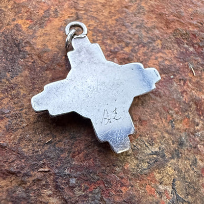 A silver-toned metal pendant shaped like a cross outline with a small loop at the top, photographed resting on a textured brown rock surface. On it's surface, a hallmark, "A. E" is scratched.
