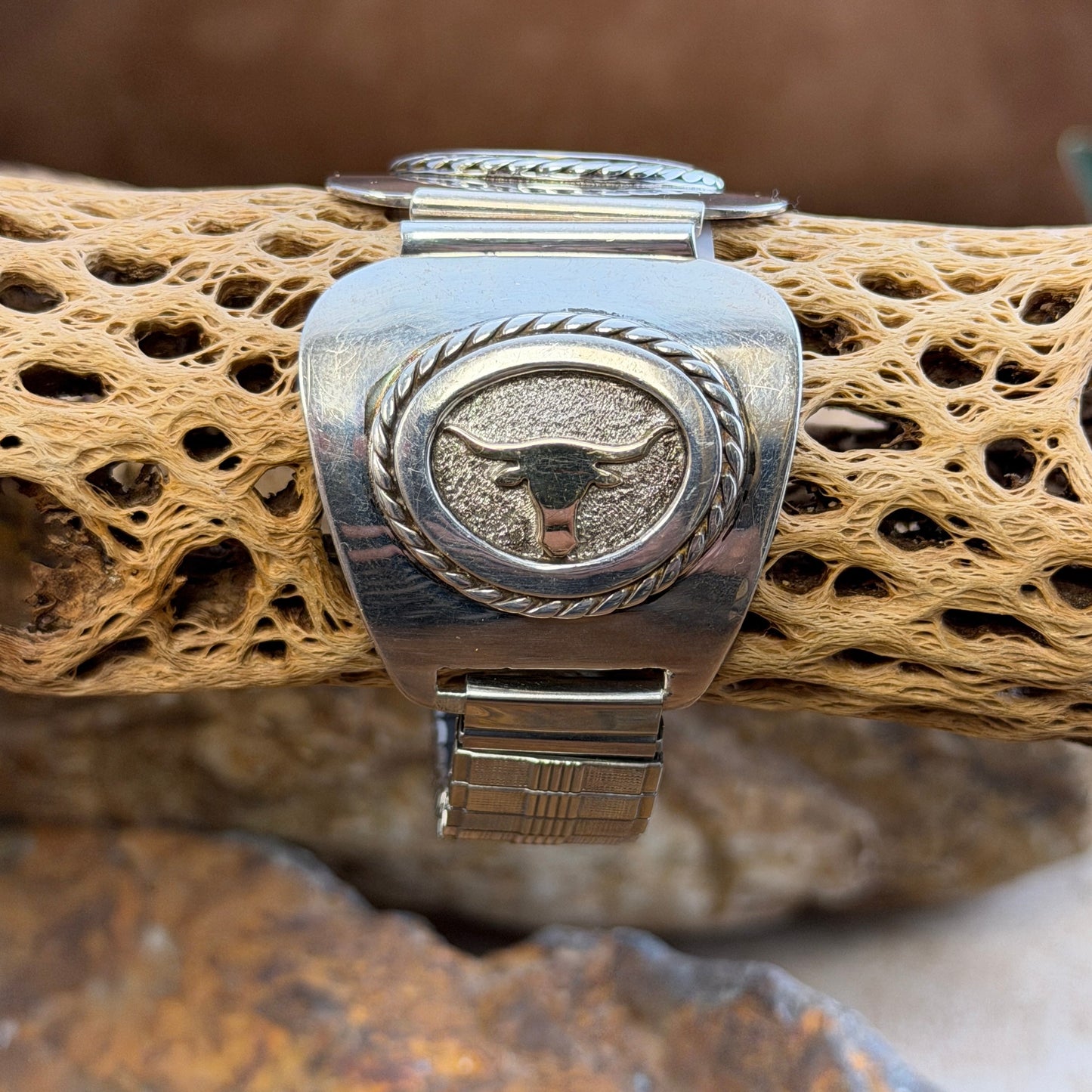 A sterling silver and 14k gold watch band decorative featuring a bull skull motif, displayed on a textured tan branch with a natural outdoor background.