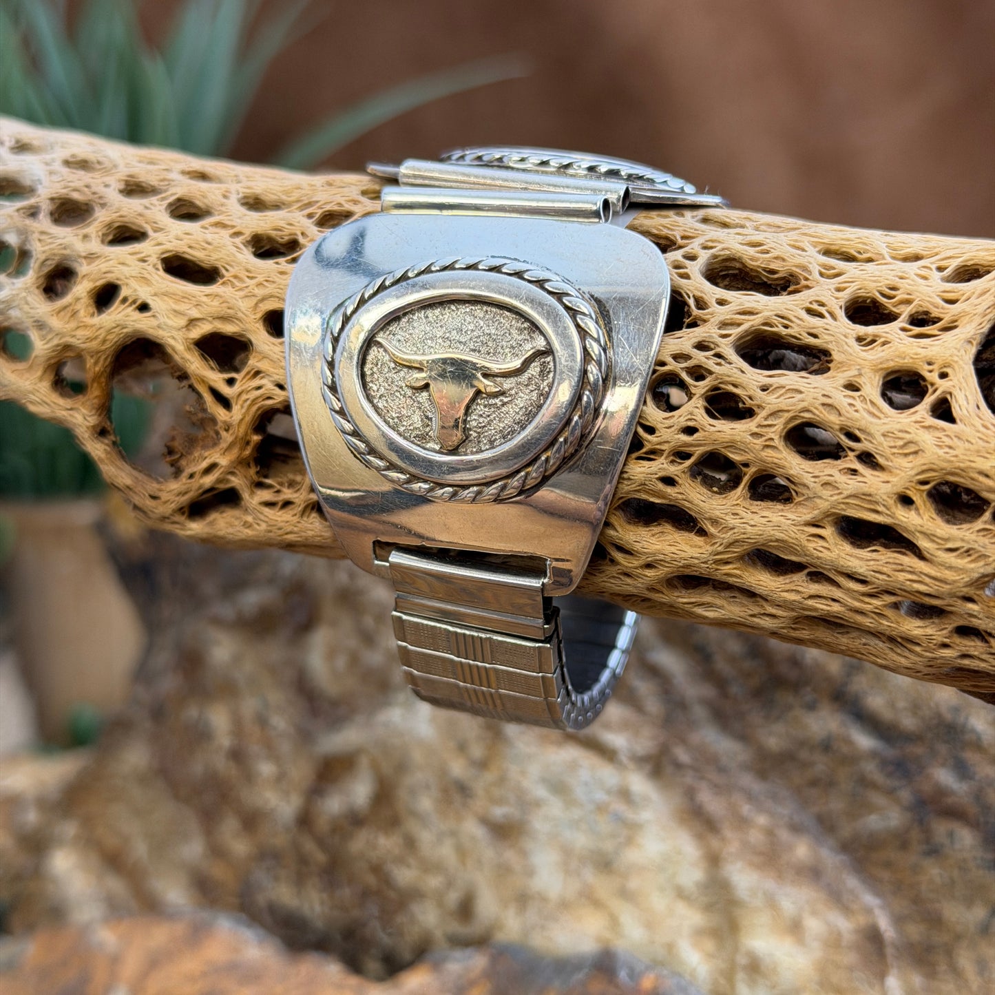 A sterling silver and 14k gold watch band decorative featuring a bull skull motif, displayed on a textured tan branch with a natural outdoor background.