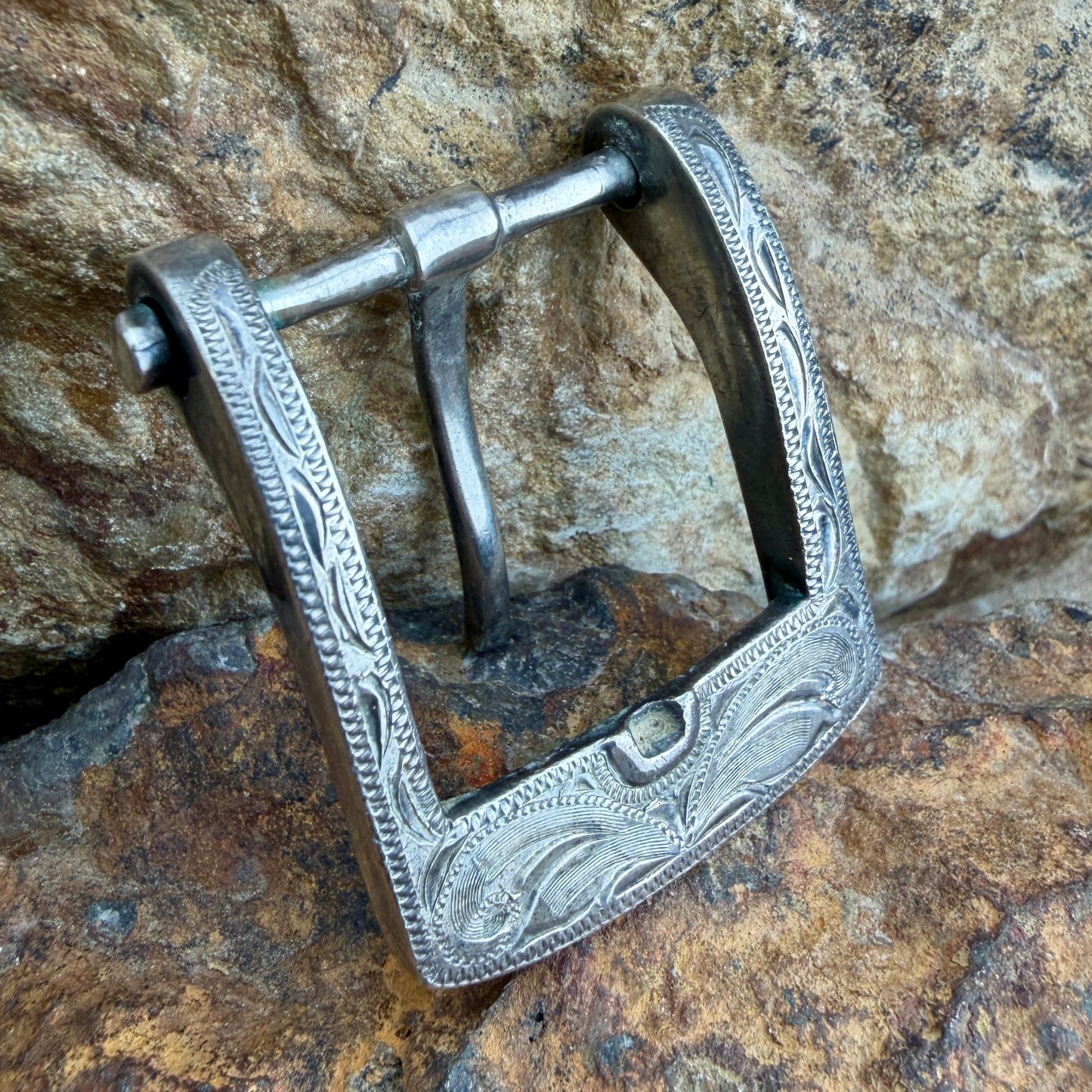 An ornate silver metal buckle with engraved floral and scrollwork patterns, shown resting on a textured brown and gray rock surface.