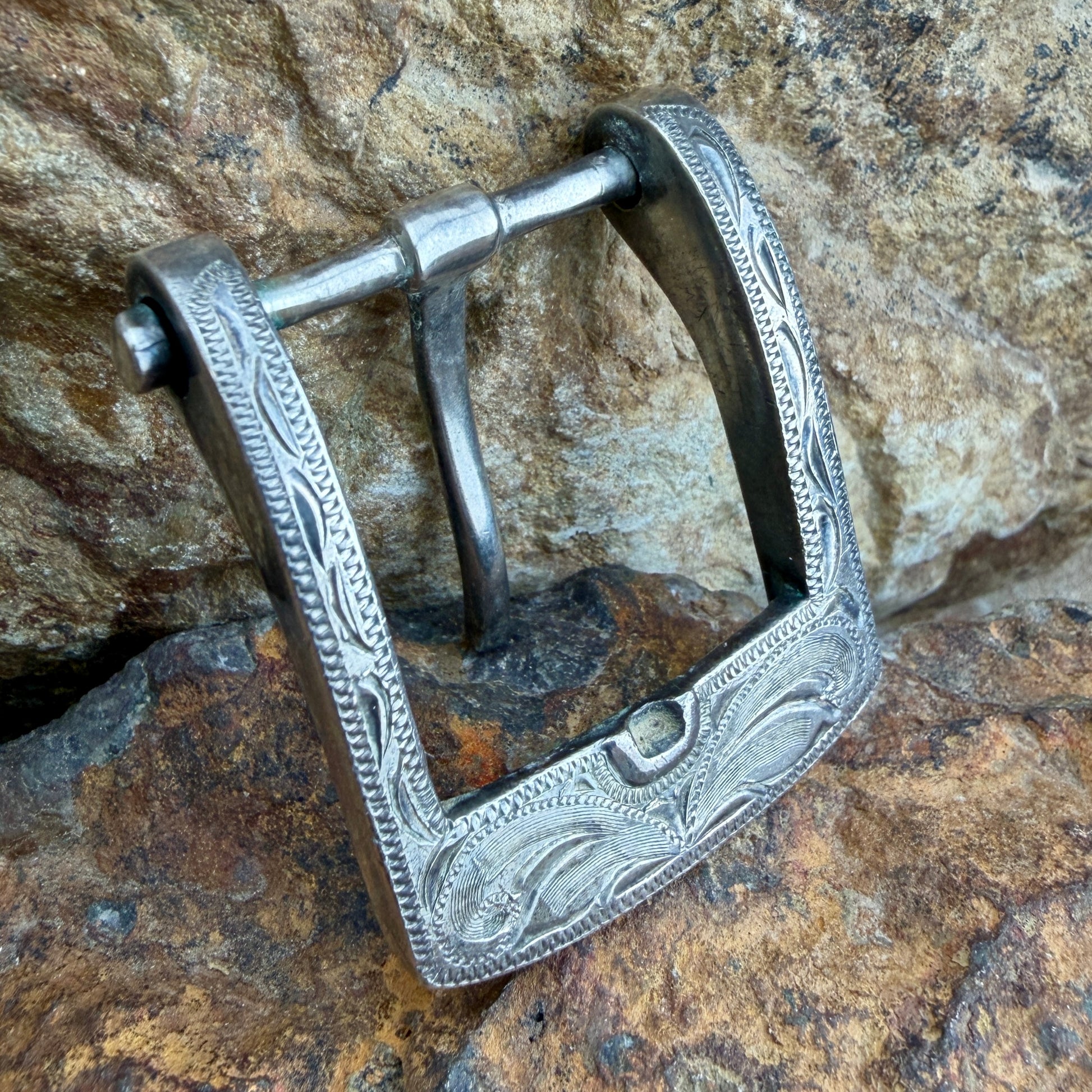 An ornate silver metal buckle with engraved floral and scrollwork patterns, shown resting on a textured brown and gray rock surface.