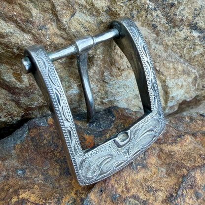 An ornate silver metal buckle with engraved floral and scrollwork patterns, shown resting on a textured brown and gray rock surface.