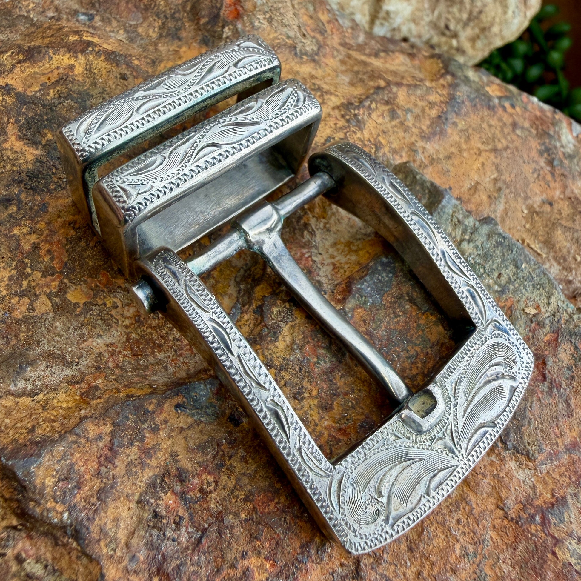 An ornate silver-toned metal belt buckle with engraved floral and geometric patterns, resting on a textured brown rock surface.