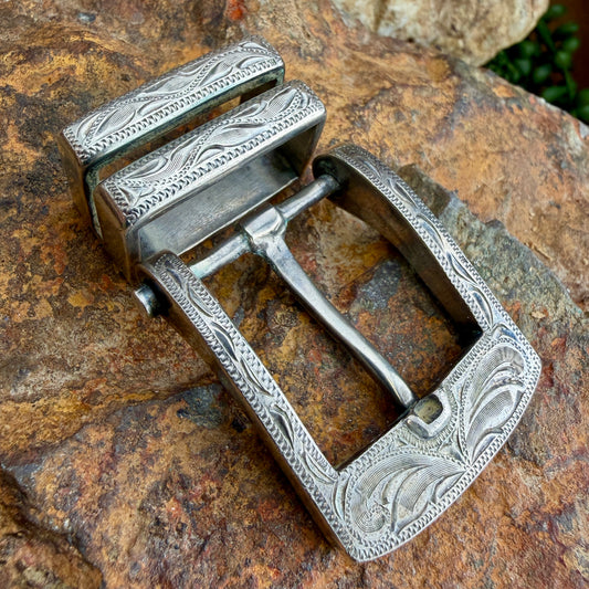 An ornate silver-toned metal belt buckle with engraved floral and geometric patterns, resting on a textured brown rock surface.