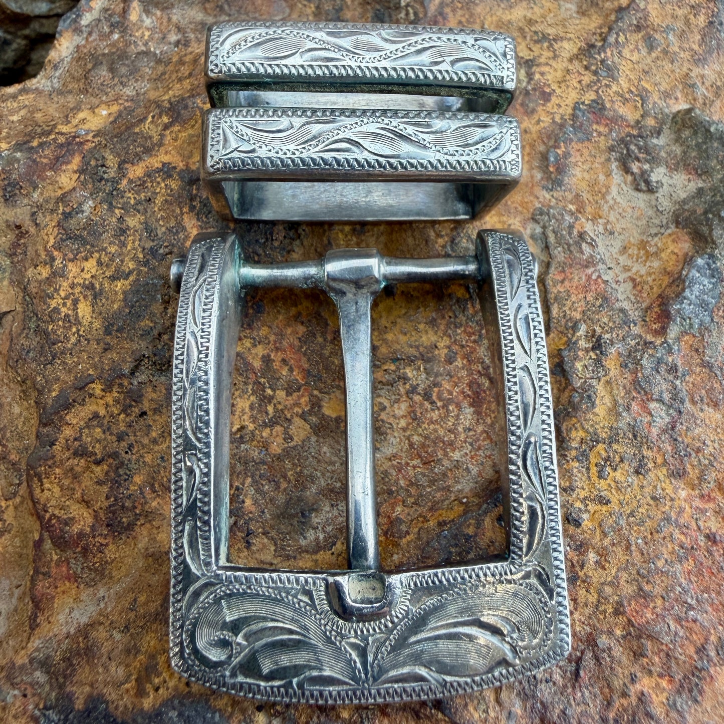 An ornate silver-toned metal belt buckle with engraved floral and scrollwork patterns, displayed on a textured brown and tan rock surface.