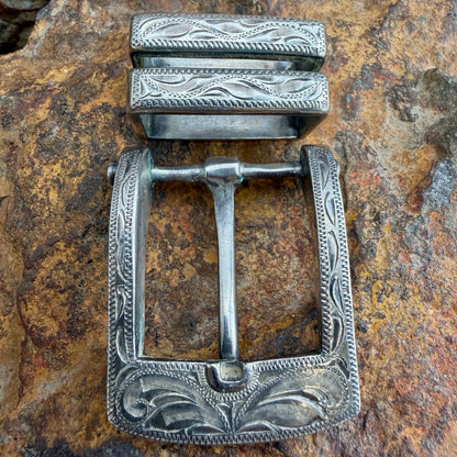 An ornate silver-toned metal belt buckle with engraved floral and scrollwork patterns, displayed on a textured brown and tan rock surface.