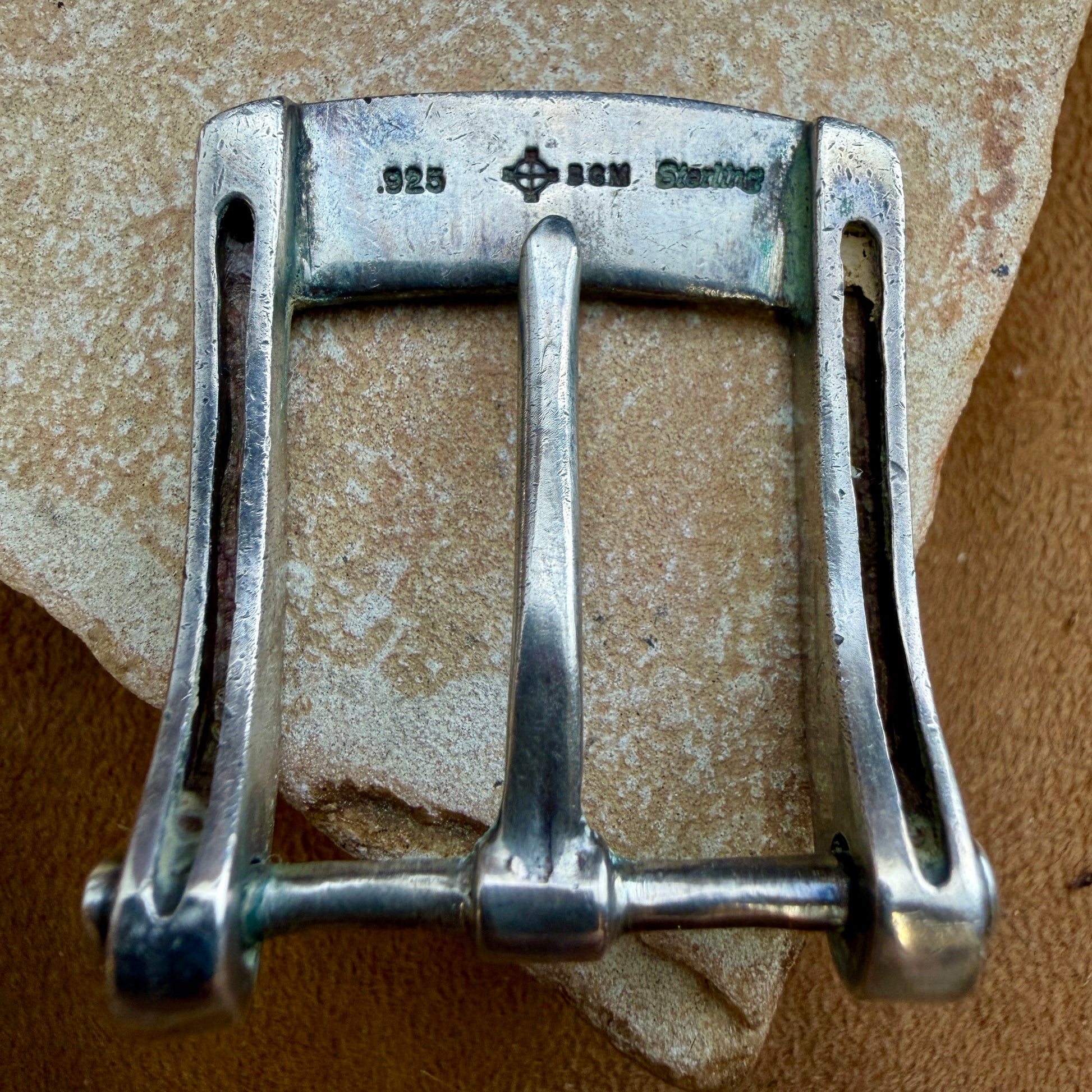 A sterling silver belt buckle with '925 BGM Sterling' engraved on the top bar, resting on a textured beige stone surface.