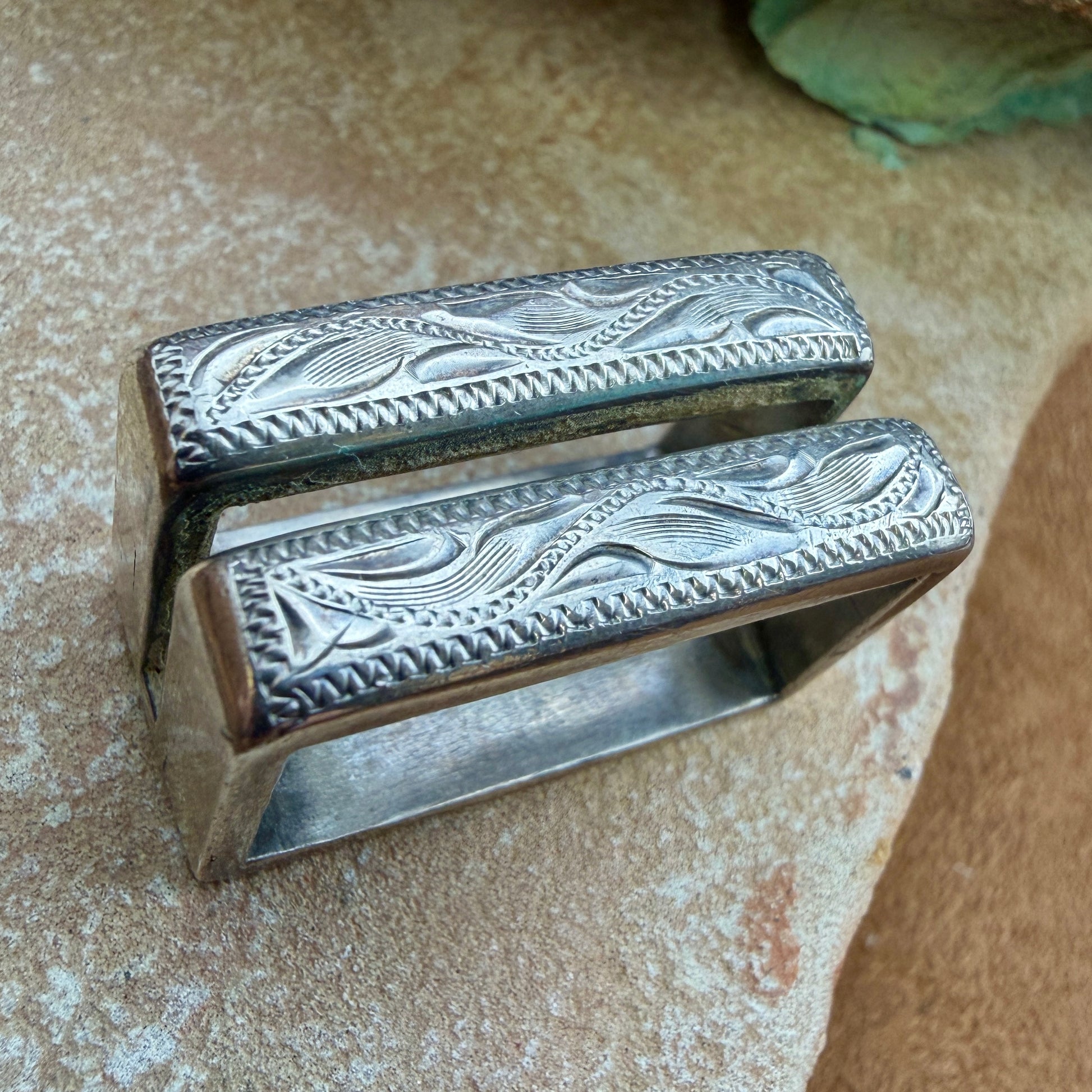 A silver-toned metal ring for a belt buckle with ornate engraved wave and leaf patterns on its rectangular band, photographed on a textured beige stone surface.