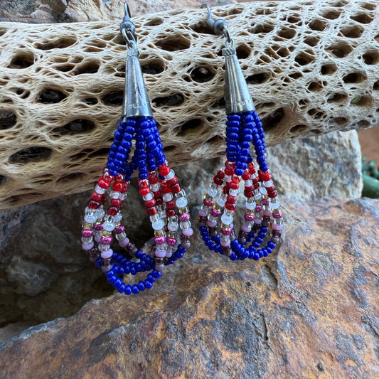 A pair of handmade beaded dangle earrings with blue, red, and white beads, hanging from silver fishhook wires against a natural rock and coral background.
