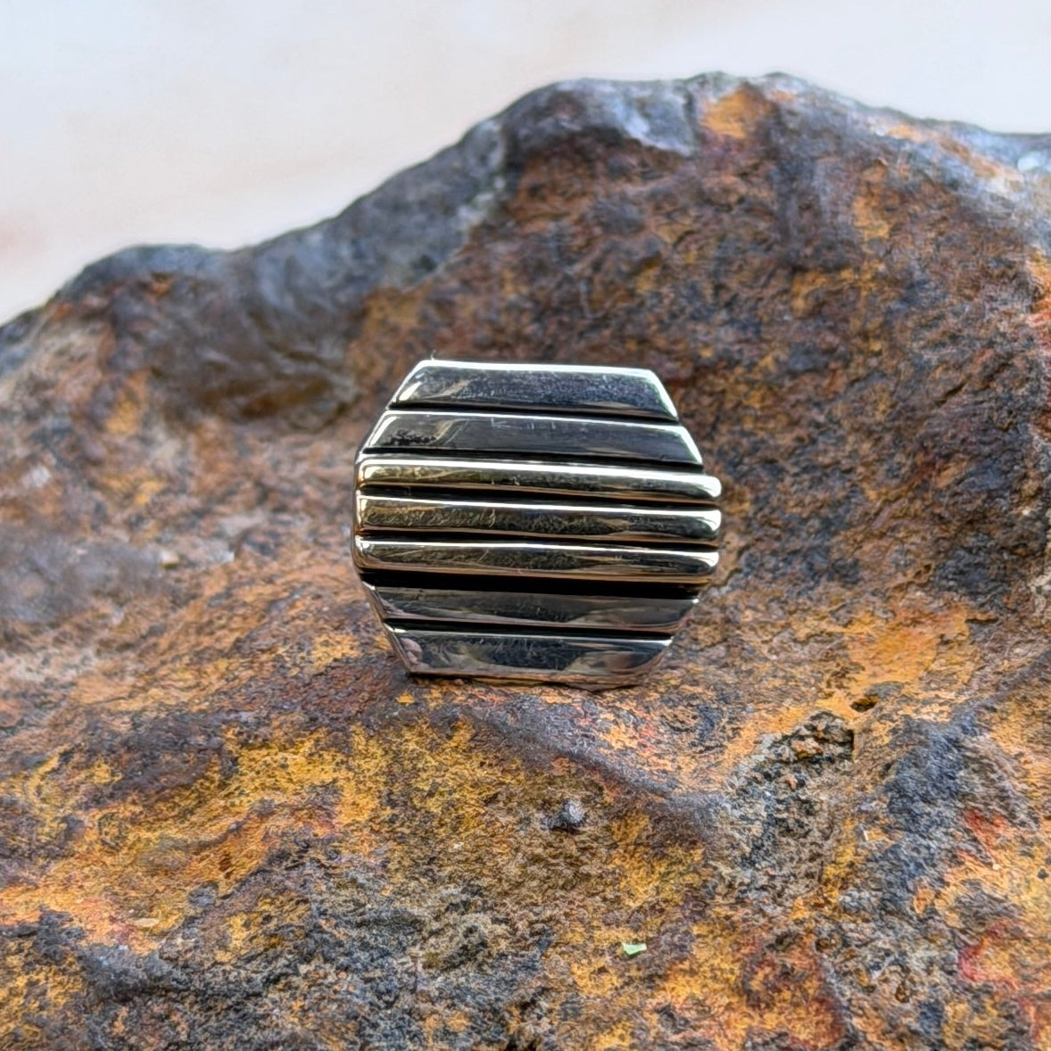 A small rectangular metal accessory with horizontal ridges sits centered on a large textured rock in an outdoor setting with blurred plants and another rock in the background.