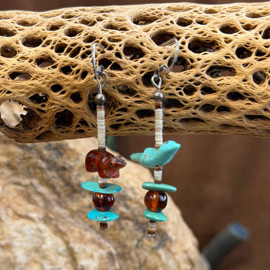 A pair of dangle earrings with cream colored heishi beads that feature two distinct animals cut from stone. on one earing a turquoise colored bird, on the other a amber colored otter. Each earring ends with a simple three item pattern of turquoise, amber, and turquoise.