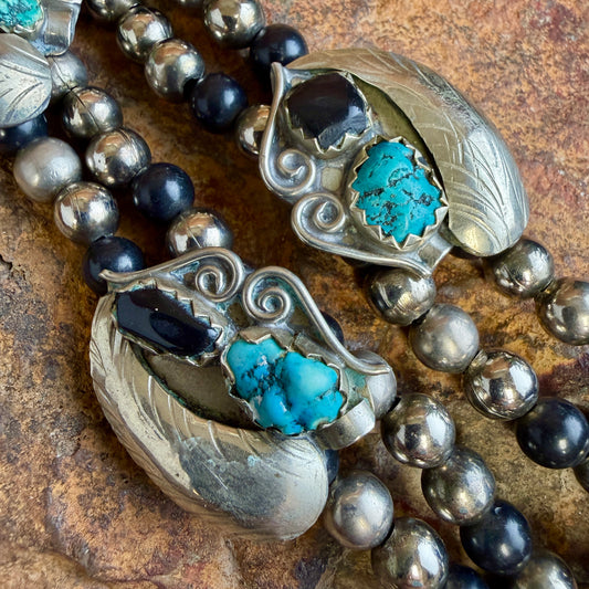 Close-up of a beaded necklace with large silver-toned leaf-shaped pendants featuring turquoise and black stone accents, layered over dark and metallic beads on a textured brown surface.