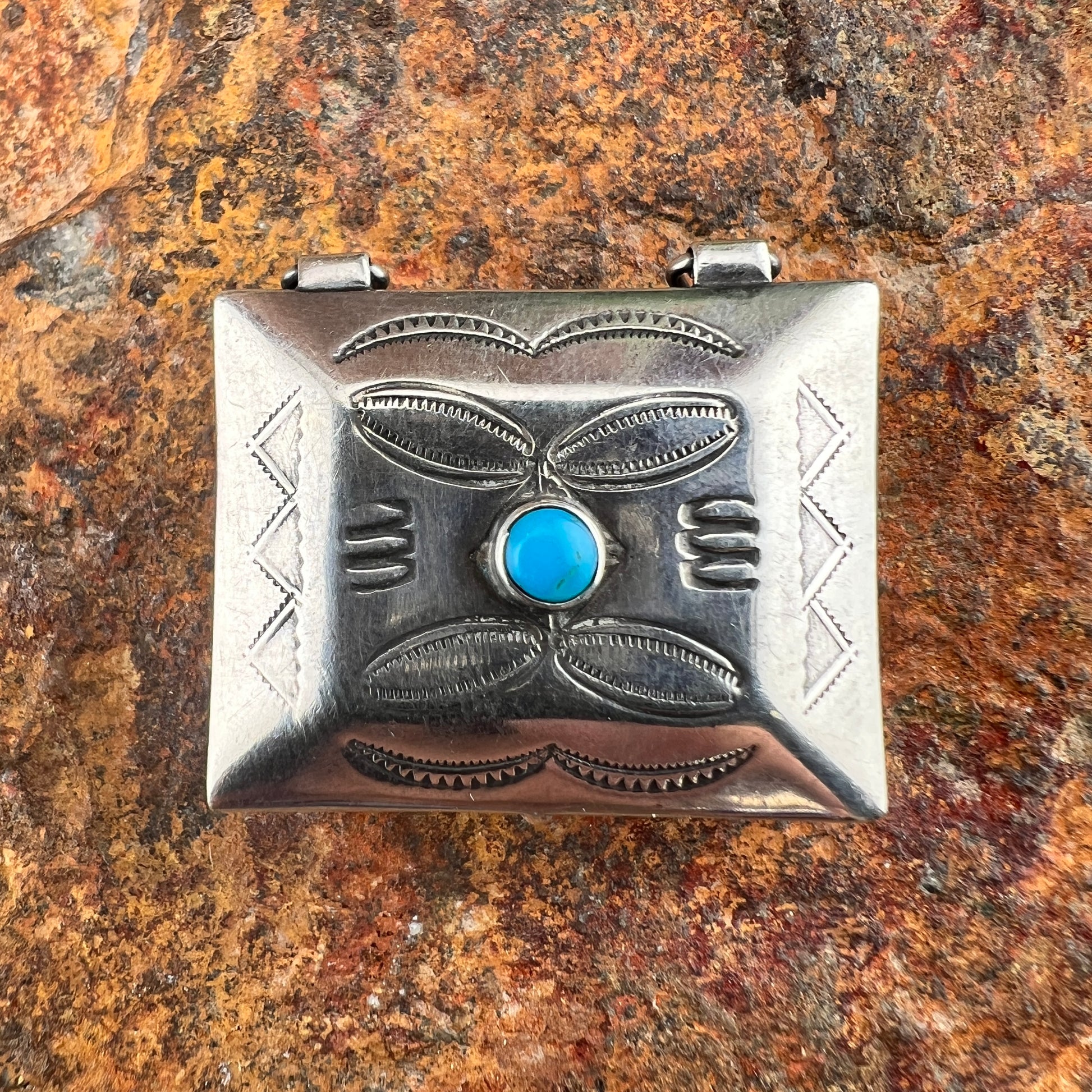 A square sterling silver jewelry box with traditional engraved patterns and a central round turquoise stone, displayed on a textured brown and orange rock surface.