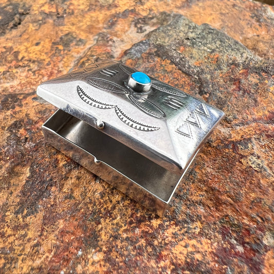A small silver-toned metal box with engraved details, featuring a round blue stone accent on the lid, photographed on a textured brown and orange rock surface.