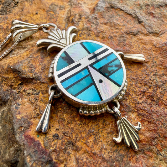 A sterling silver pendant representing a Yei face. It is inlaid with various shades of blue and black stones. Attached are silver fanned accents that hang from three anchor points. 