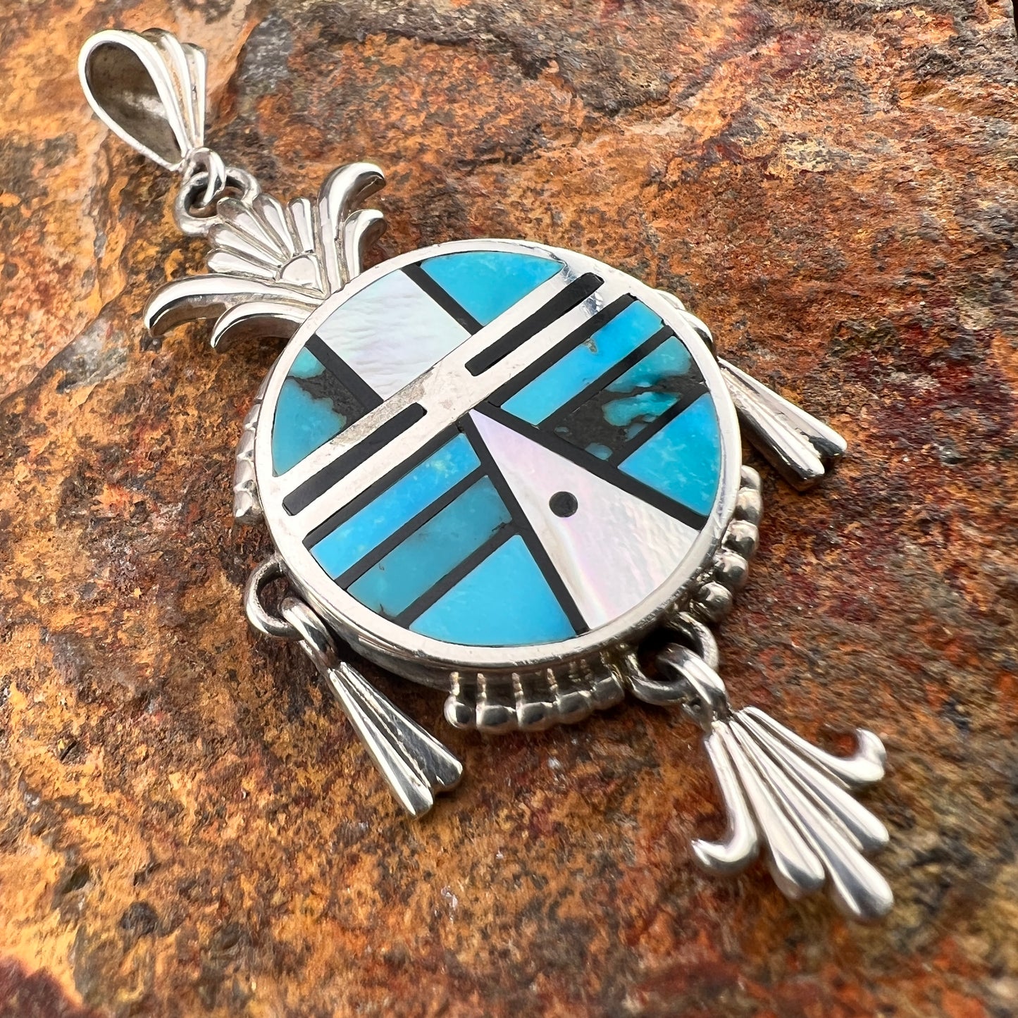 A sterling silver pendant representing a Yei face. It is inlaid with various shades of blue and black stones. Attached are silver fanned accents that hang from three anchor points. 