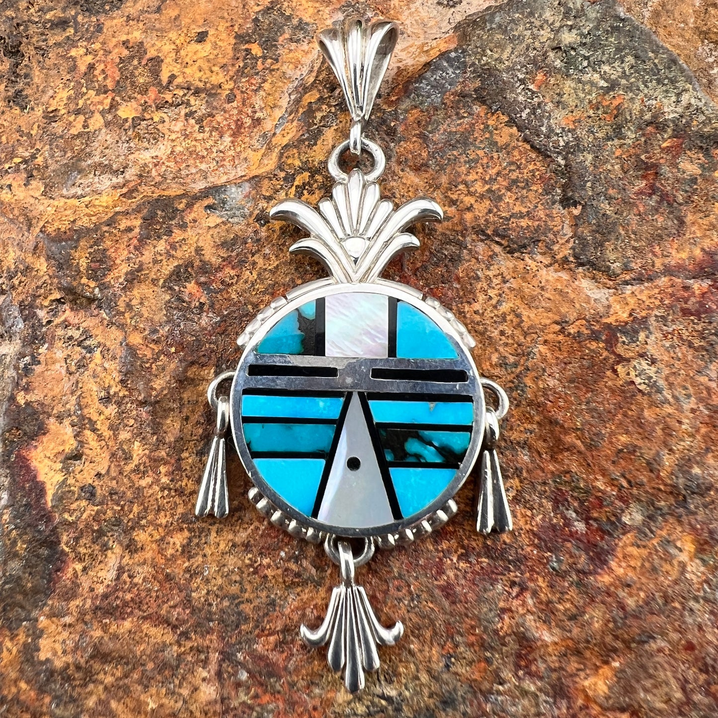 A sterling silver pendant representing a Yei face. It is inlaid with various shades of blue and black stones. Attached are silver fanned accents that hang from three anchor points. 