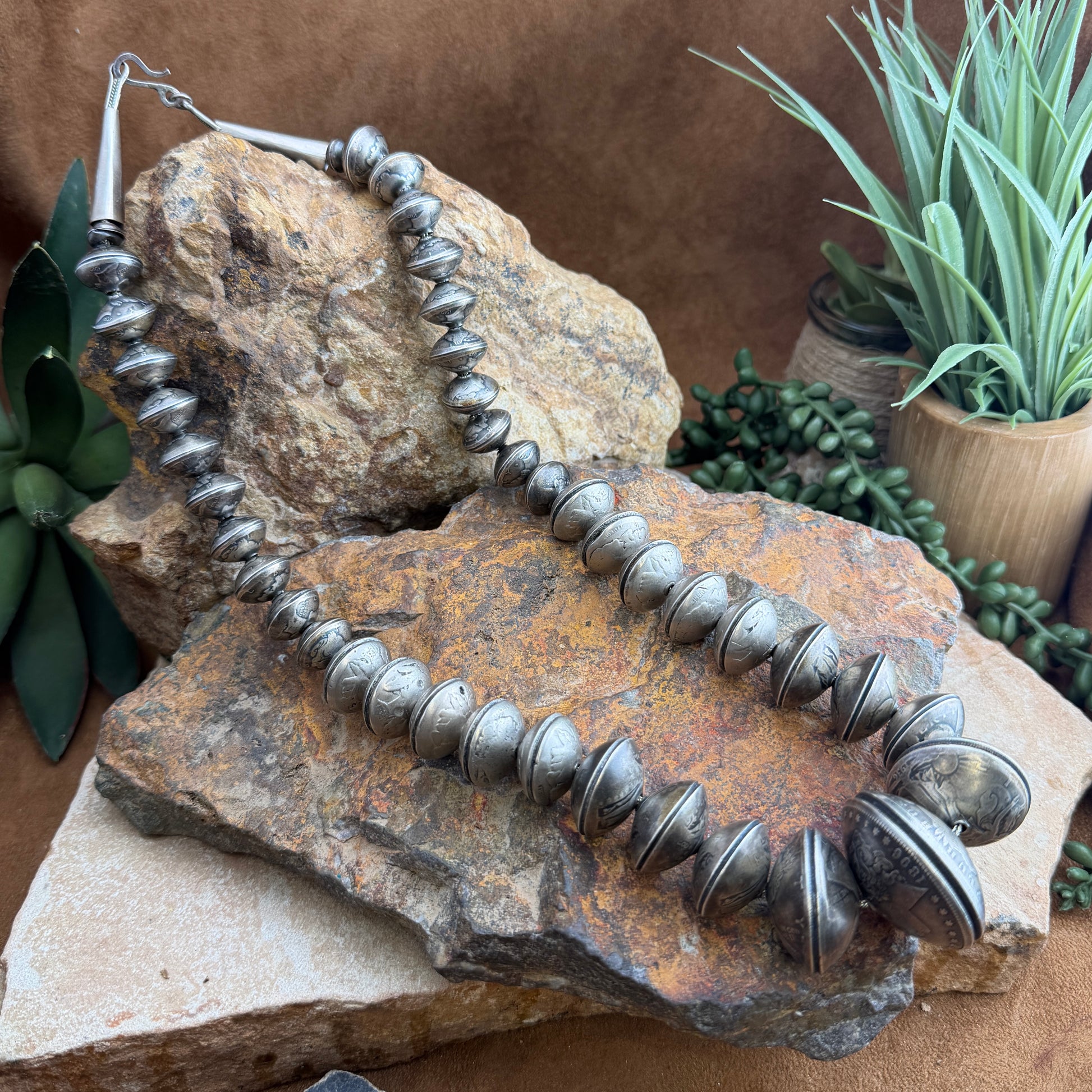 Silver beaded necklace made of coins on a textured stone surface with plants in the background