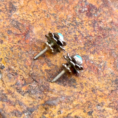 A pair of small floral stud earrings with oxidized bronze metal petals and light blue turquoise stones, shown on a textured brown and orange stone surface.
