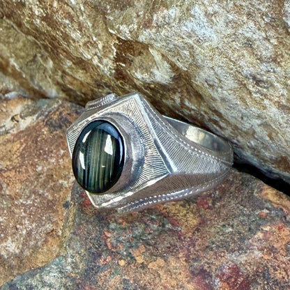 A silver-toned textured ring with a large oval black stone, photographed resting on rough natural rocks.