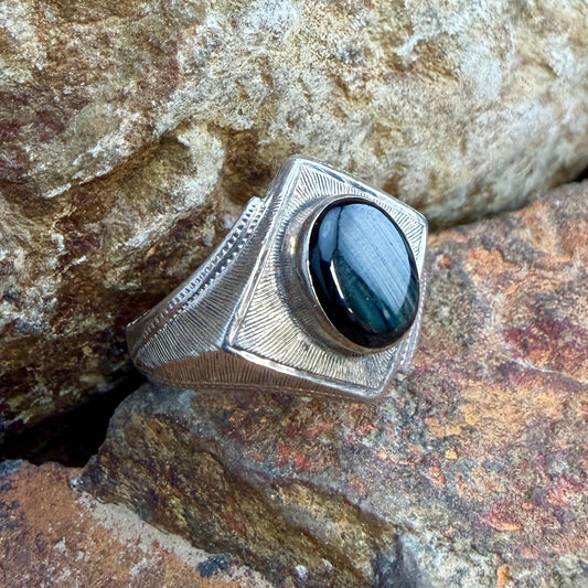 A silver-toned linear textured ring with a large oval black stone, photographed resting on rough natural rocks.