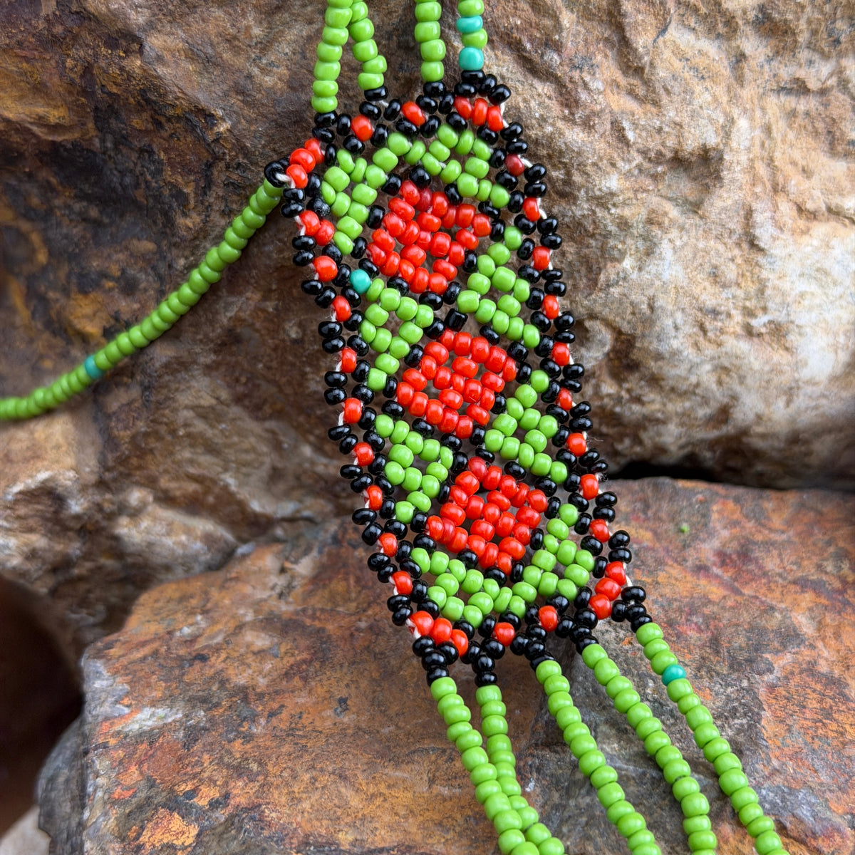 Close up of a the beaded bracelet with red, green, and black seed beads, displayed on a textured brown rock background.