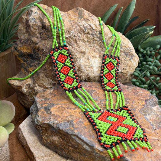 A beaded necklaces with geometric patterns in green, red, and black, displayed on natural rocks with succulent plants.