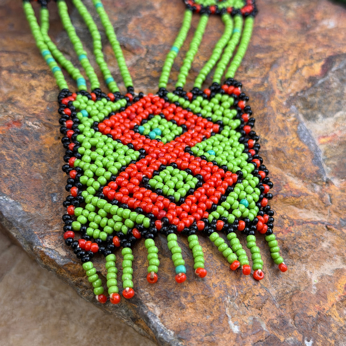 A square beaded necklace with a bold geometric pattern in red, lime green, black, and small turquoise accents, displayed on a textured brown stone surface.
