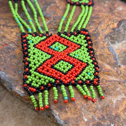 A square beaded necklace with a bold geometric pattern in red, lime green, black, and small turquoise accents, displayed on a textured brown stone surface.