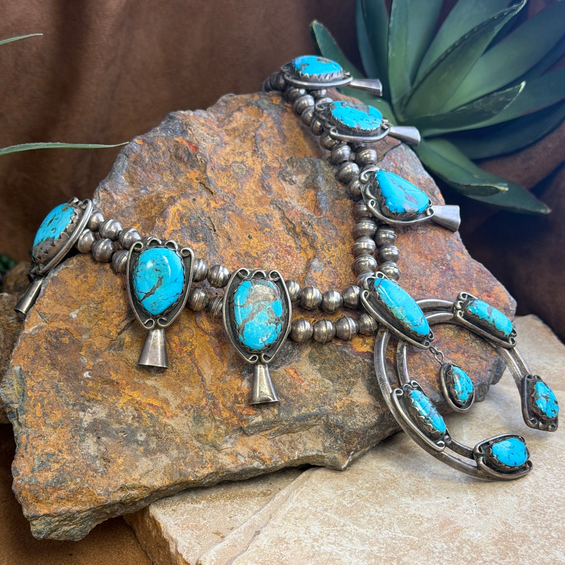 A silver-toned Southwestern-style necklace with multiple turquoise stones, draped over a large textured brown rock with a green succulent in the background.