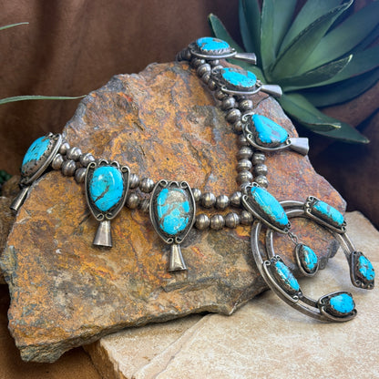 A silver-toned Southwestern-style necklace with multiple turquoise stones, draped over a large textured brown rock with a green succulent in the background.
