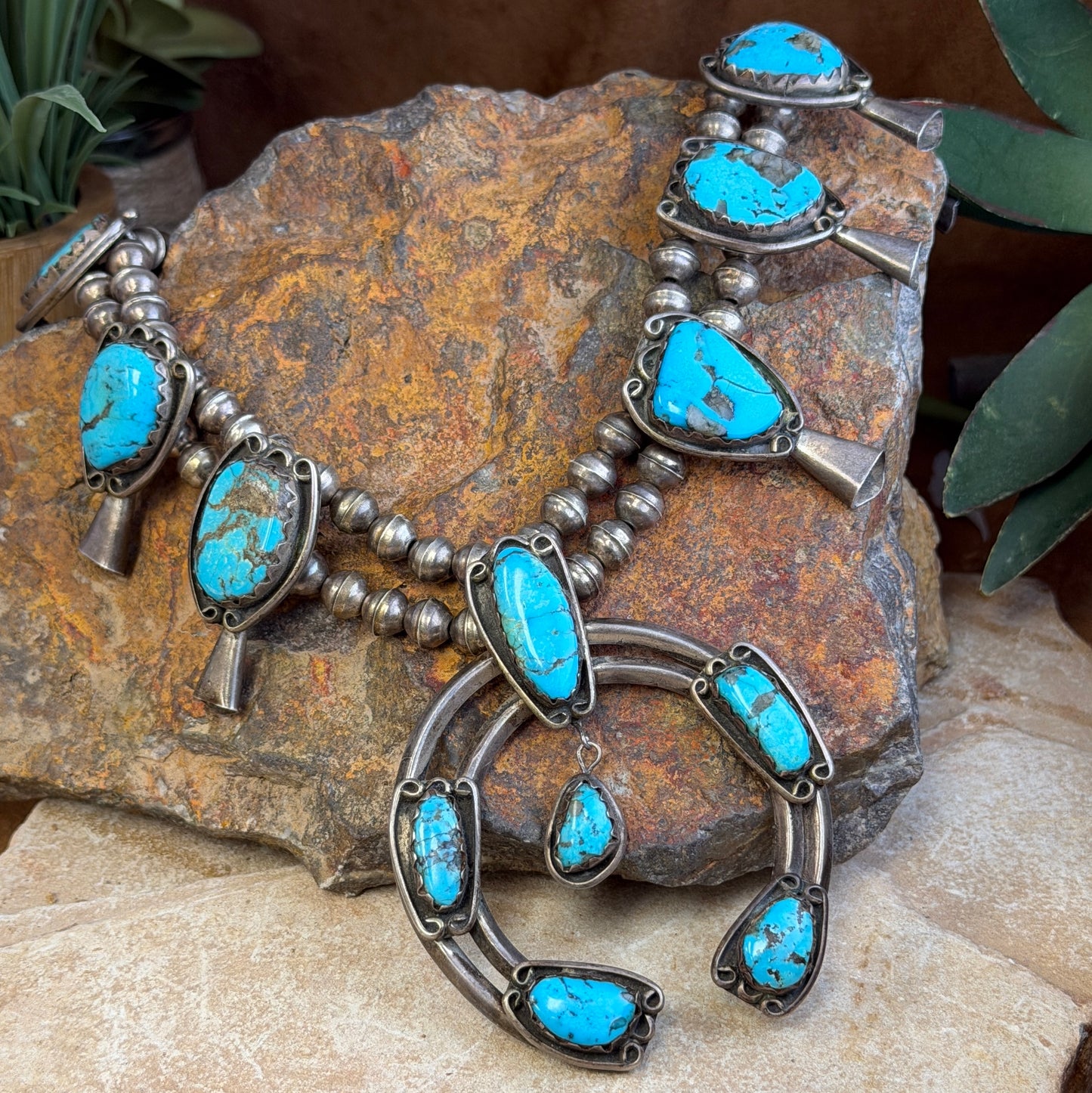 A Southwestern-style turquoise and silver necklace displayed on a textured brown rock with green leaves in the background.