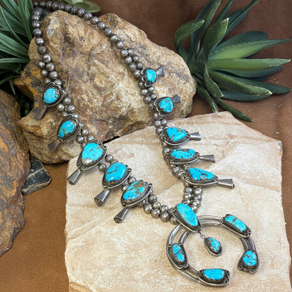 A silver-toned Southwestern-style turquoise necklace displayed on a stone slab with decorative rocks and green plants in the background.