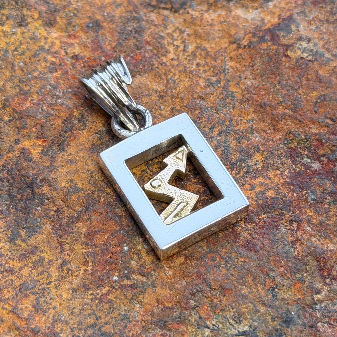 A silver square pendant with a cutout design showing a gold zig zag arrow inside, resting on a textured brown and orange rock surface.