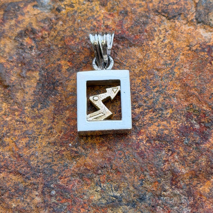 A silver-toned square pendant necklace with a gold-toned zigzag symbol inside, displayed on a textured rust-colored rock.