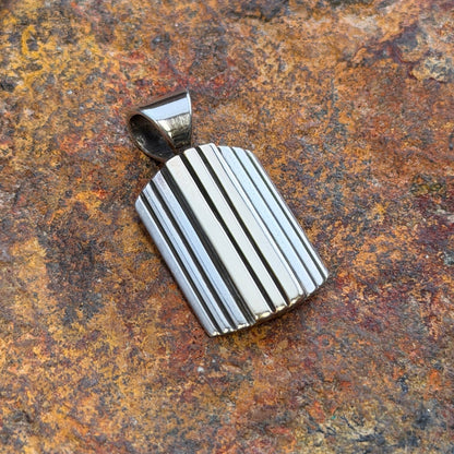 A silver-toned rectangular pendant with vertical ridges, resting on a textured orange-brown rock surface.
