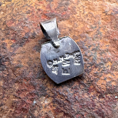 the back of a small silver pendant with engraved characters, photographed resting on a textured rusted rock surface.
