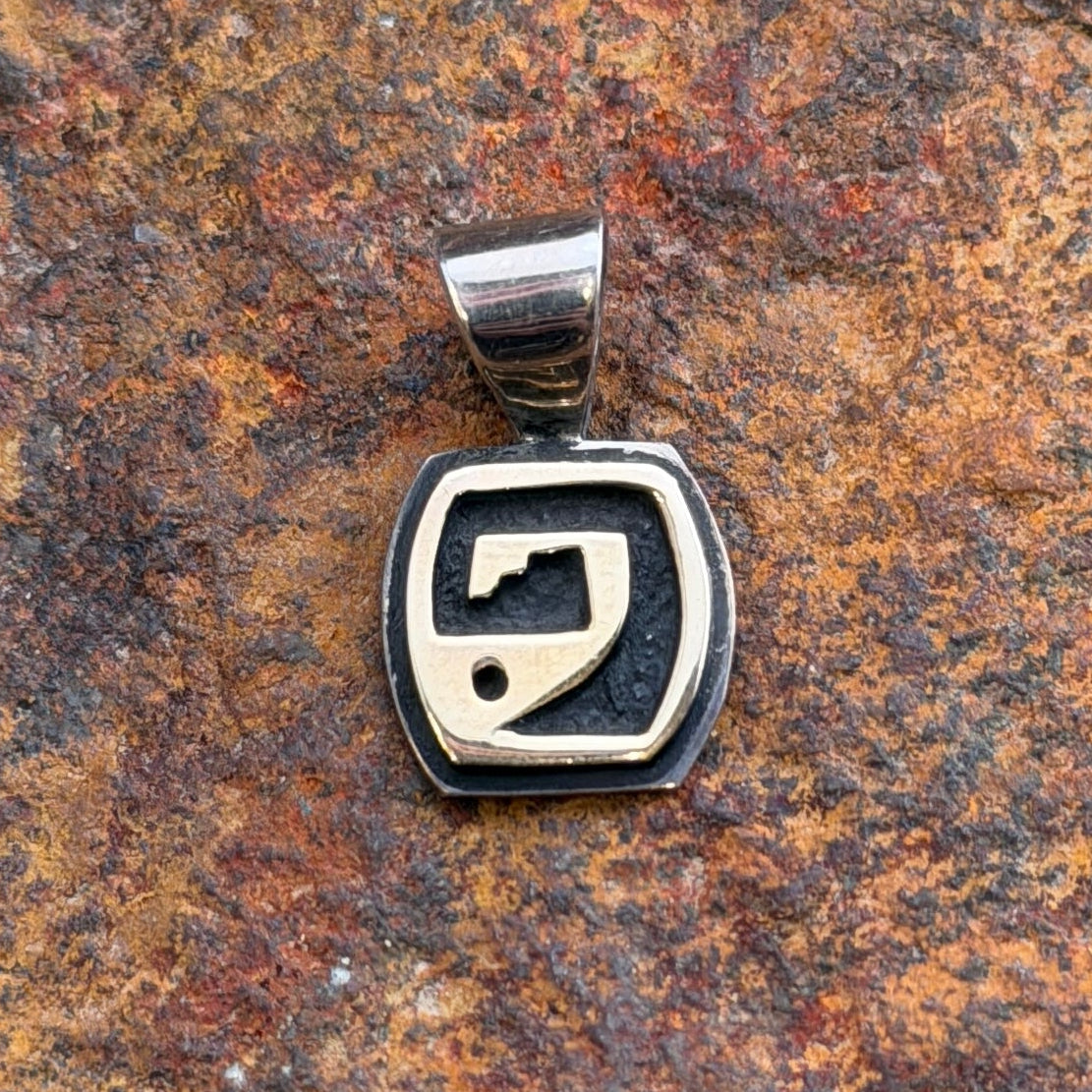 a sterling silver pendant with a gold overlayed design on a oxidized dark sterling silver pendant. it rests on an orange rust colored rock.
