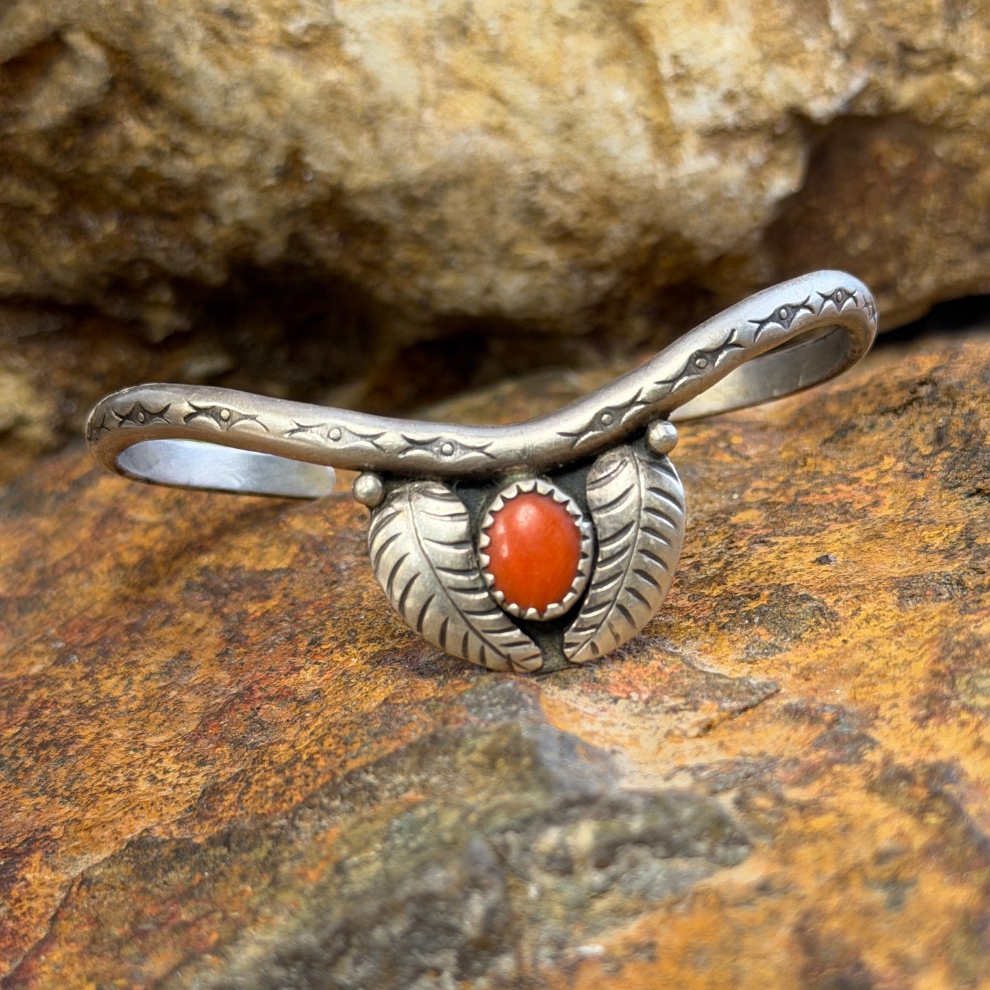 thin sterling silver cuff with a centerpiece of coral surrounded by two feathers. it is sitting on a rock surface.