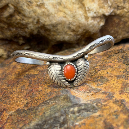 thin sterling silver cuff with a centerpiece of coral surrounded by two feathers. it is sitting on a rock surface.