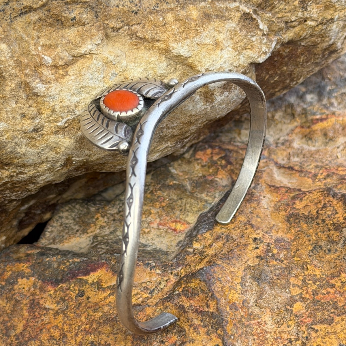 thin sterling silver cuff with a centerpiece of coral surrounded by two feathers. it is sitting on a rock surface.
