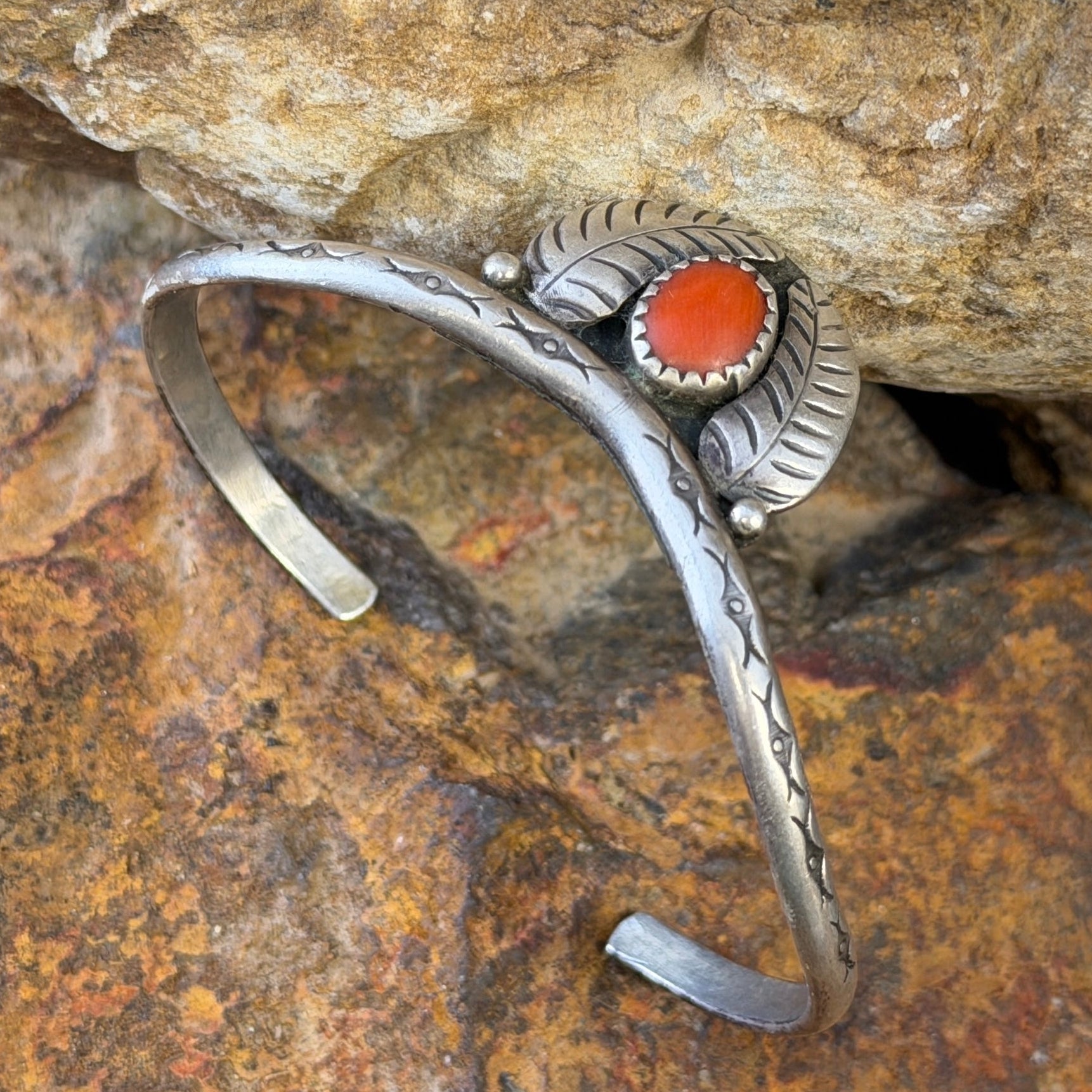 thin sterling silver cuff with a centerpiece of coral surrounded by two feathers. it is sitting on a rock surface.
