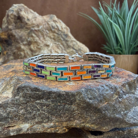 A silver-toned link bracelet with colorful rectangular enamel inlays in orange, blue, green, and purple, displayed on a textured rock with a blurred plant in the background.