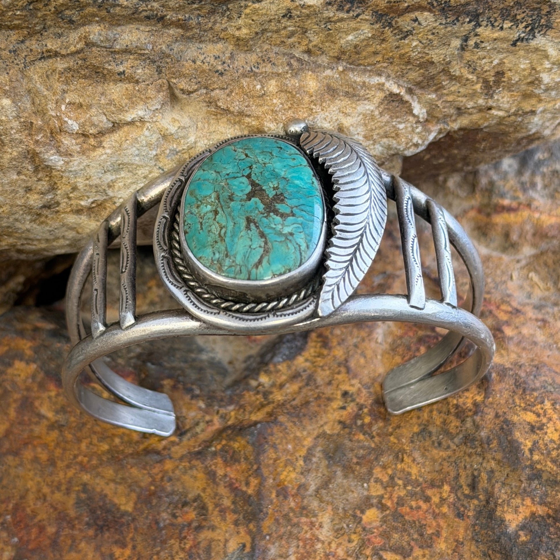 sterling silver cuff with large turquoise cabochon set in bezel surrounded by rope twist and other ornate details. it is sitting on a rock.