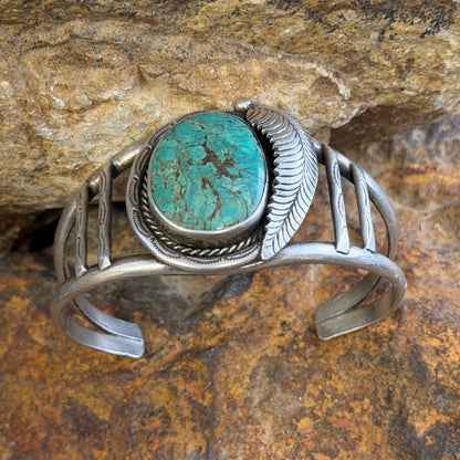 sterling silver cuff with large turquoise cabochon set in bezel surrounded by rope twist and other ornate details. it is sitting on a rock.