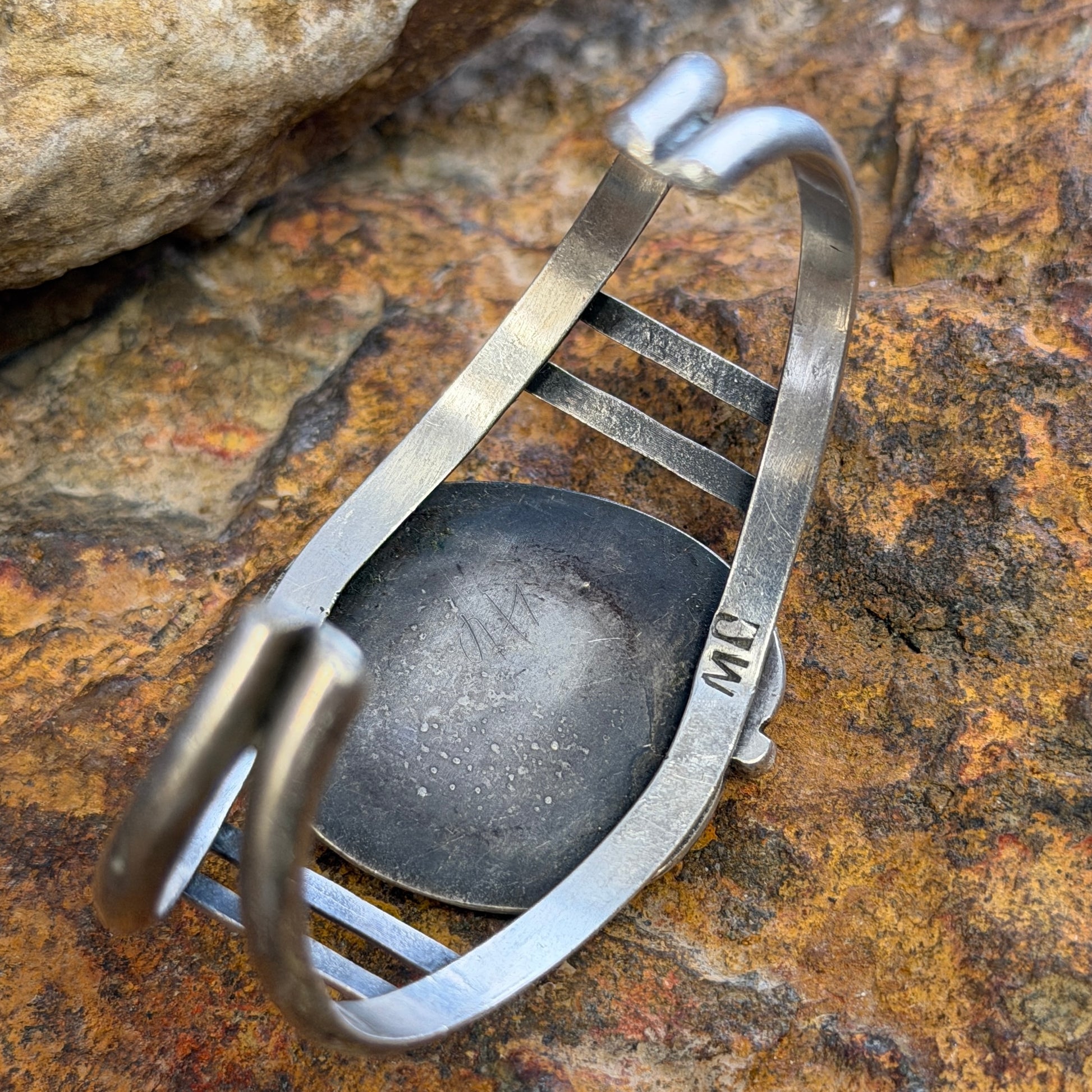 Inside of Sterling silver cuff showcasing stamping of artist "MC" it is sitting on a rock. 