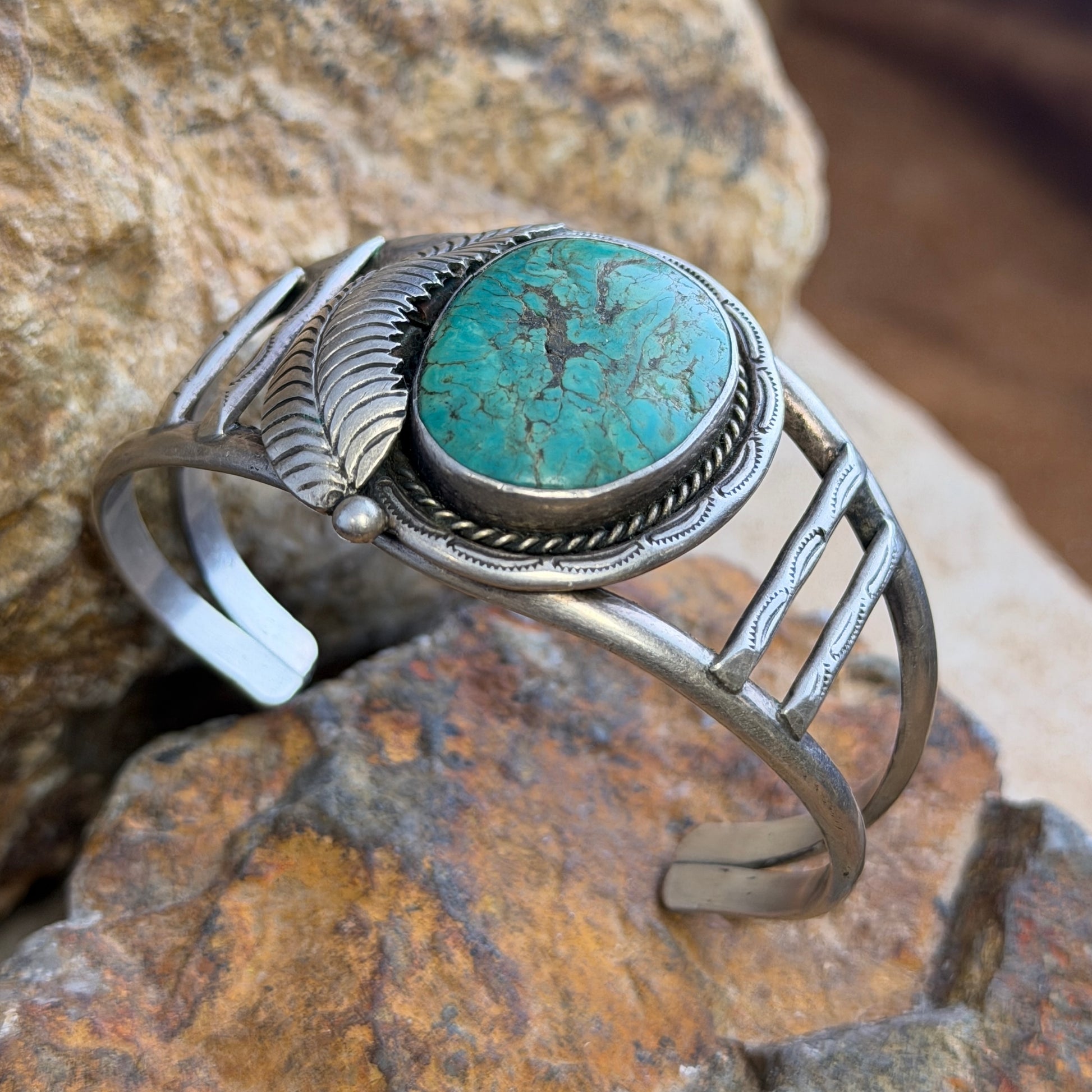 sterling silver cuff with large turquoise cabochon set in bezel surrounded by rope twist and other ornate details. it is sitting on a rock.